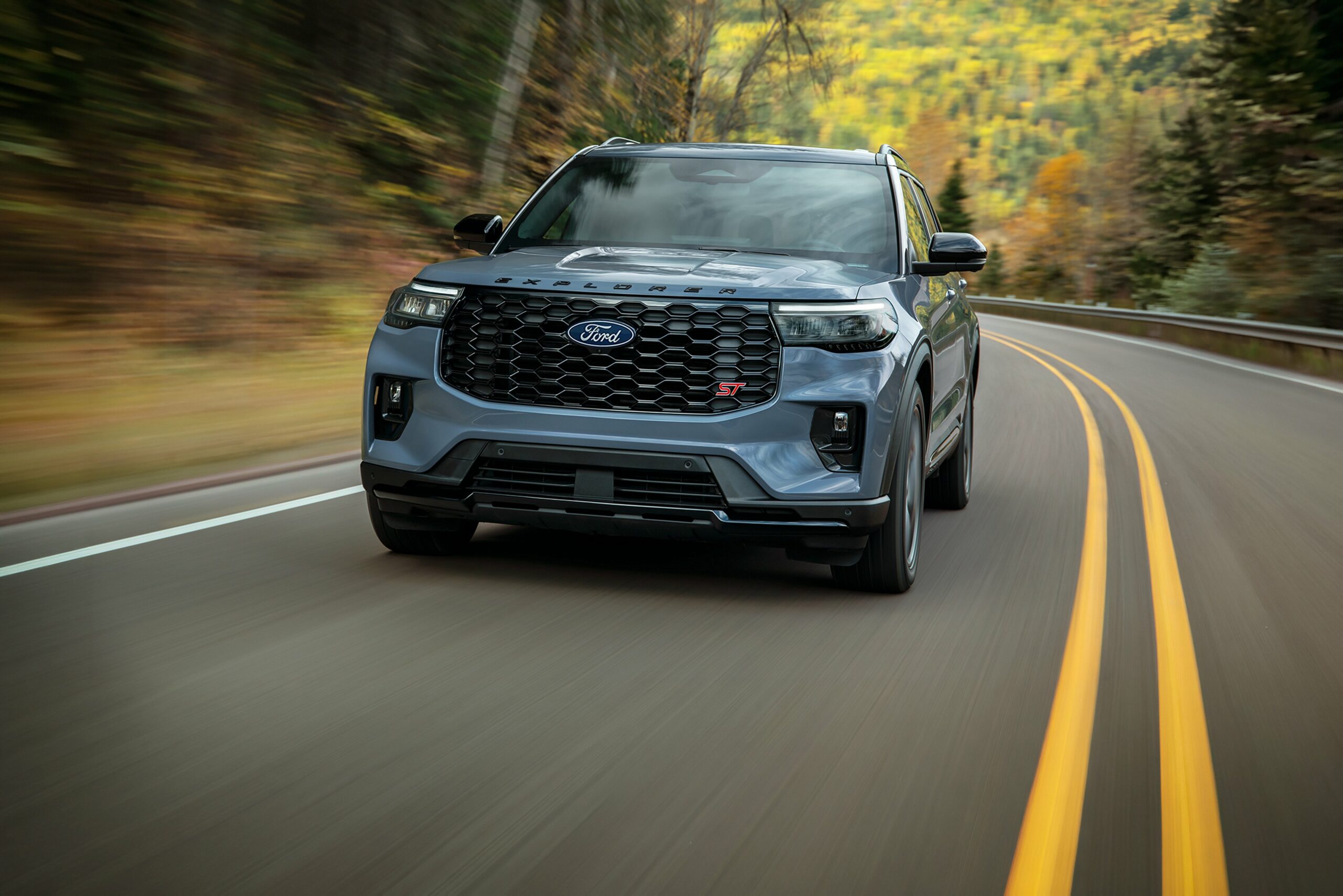 A light blue 2027 Ford Explorer driving on a curving road lined with fall foliage
