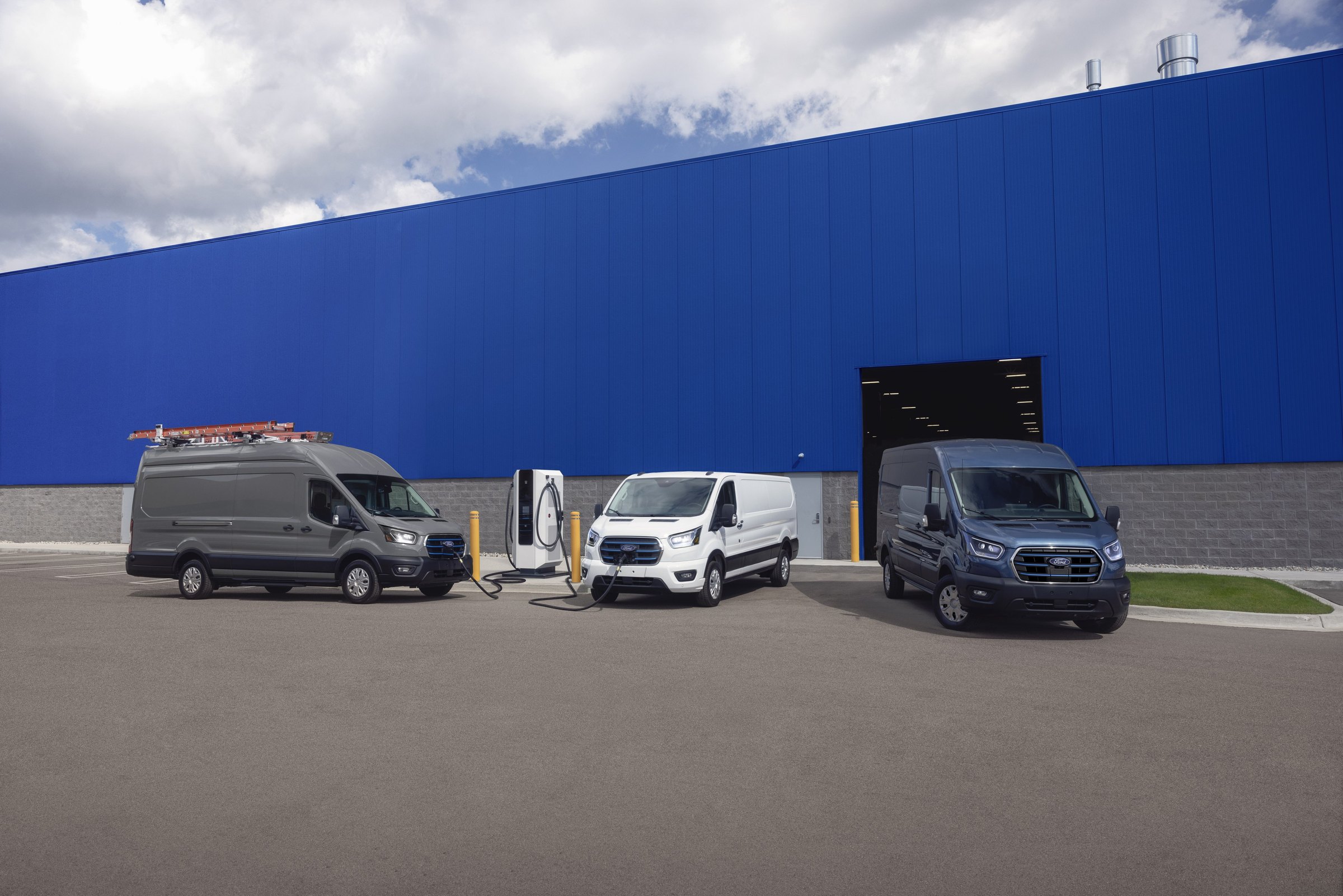 Three Ford E-Transit vans lined up outside a blue warehouse wall, the high-roof van on the left plugged into a charging station