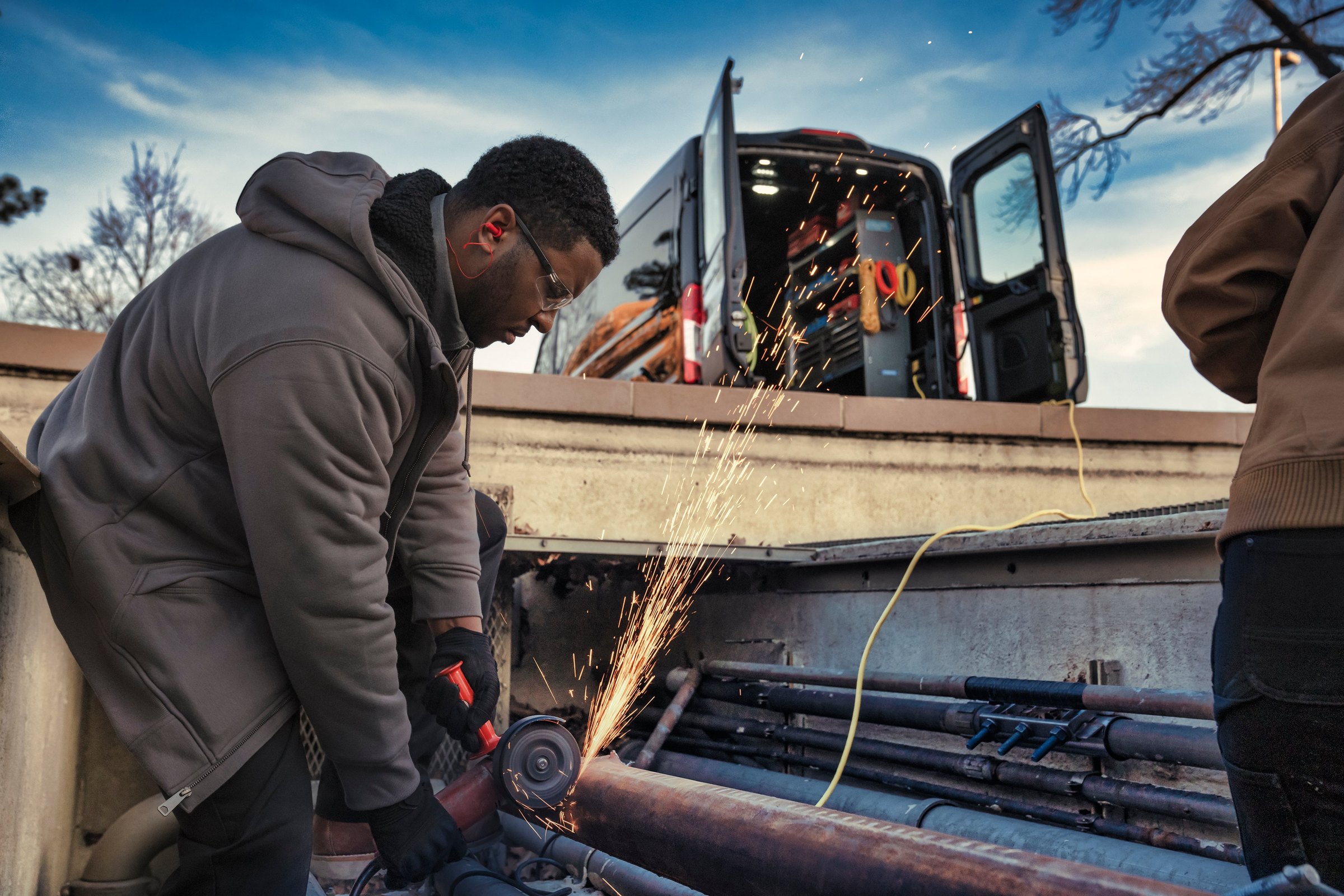 Two workers at the open rear doors of a dark Ford E-Transit at dusk, one cutting metal with an angle grinder and throwing sparks across the load floor