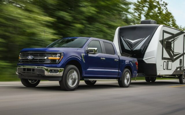 A dark blue 2026 Ford F-150 SuperCrew towing a white travel trailer along a highway
