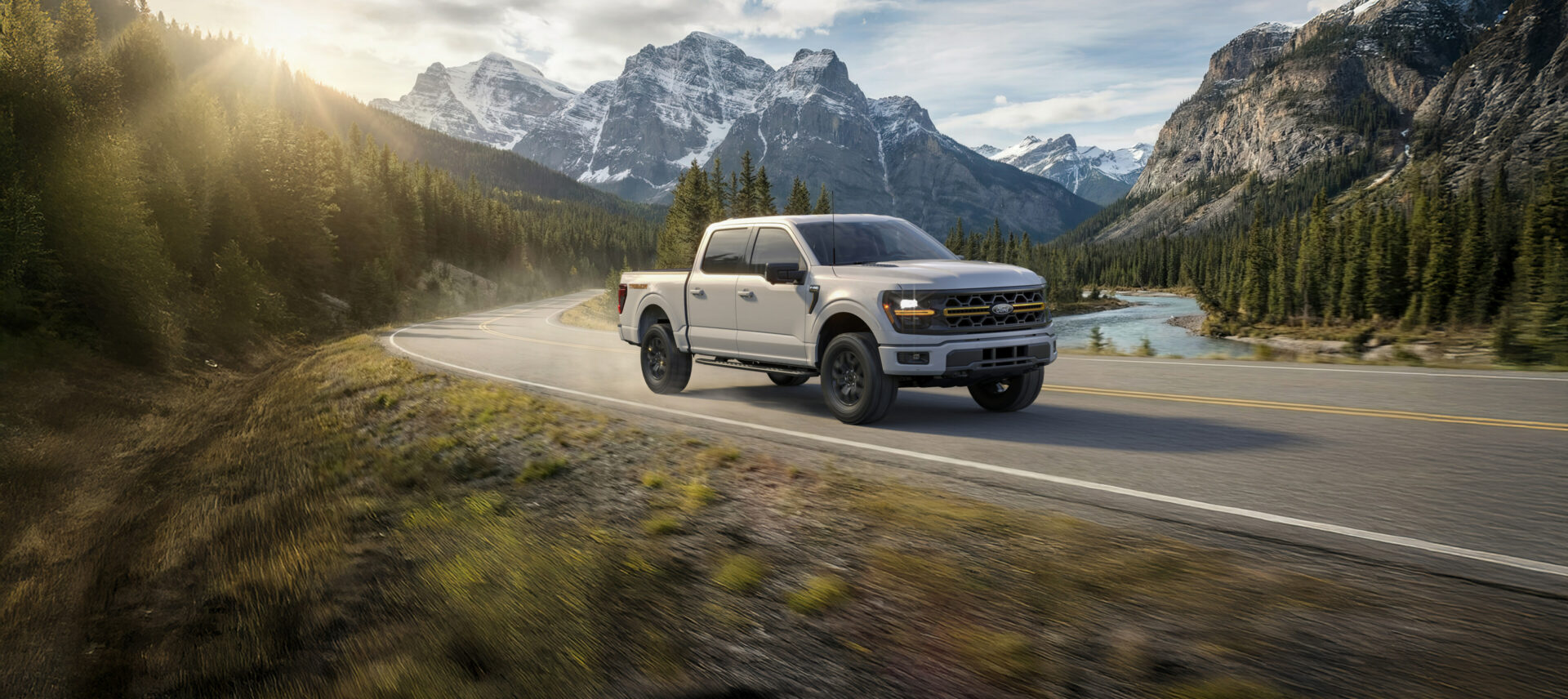 A white 2026 Ford F-150 Tremor on a curving mountain road beside a river, snow-capped peaks behind
