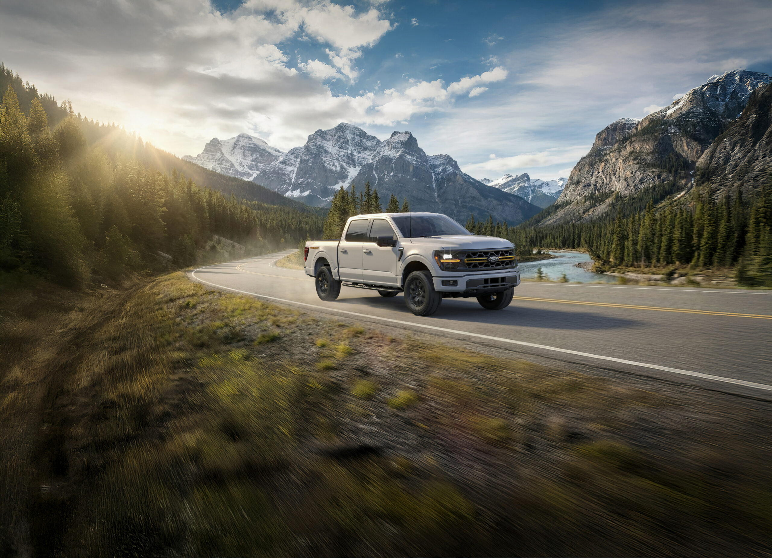A white 2026 Ford F-150 Tremor on a curving mountain road beside a river, snow-capped peaks behind