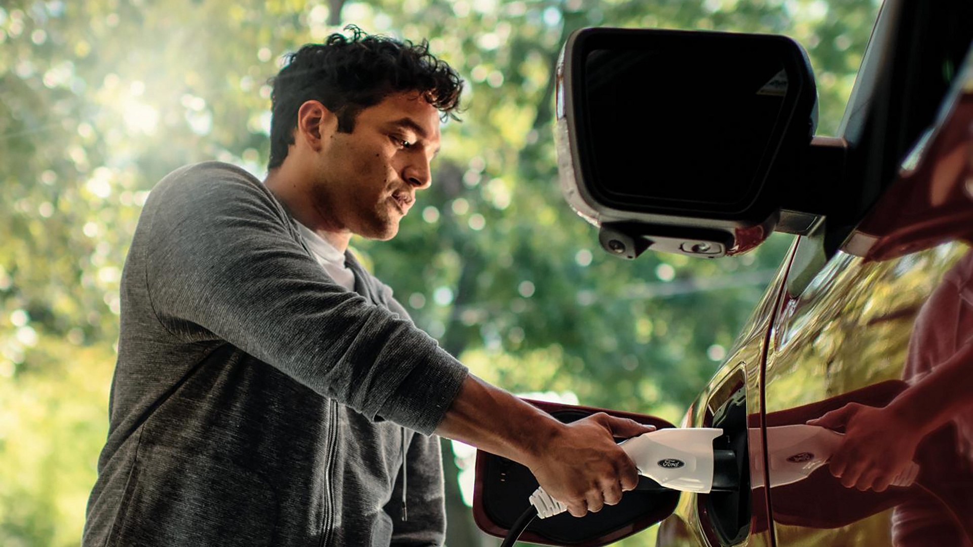 A man in a gray sweatshirt pushing a white Ford charge connector into the charge port of a red Ford F-150 Lightning, outdoors in dappled daylight under trees