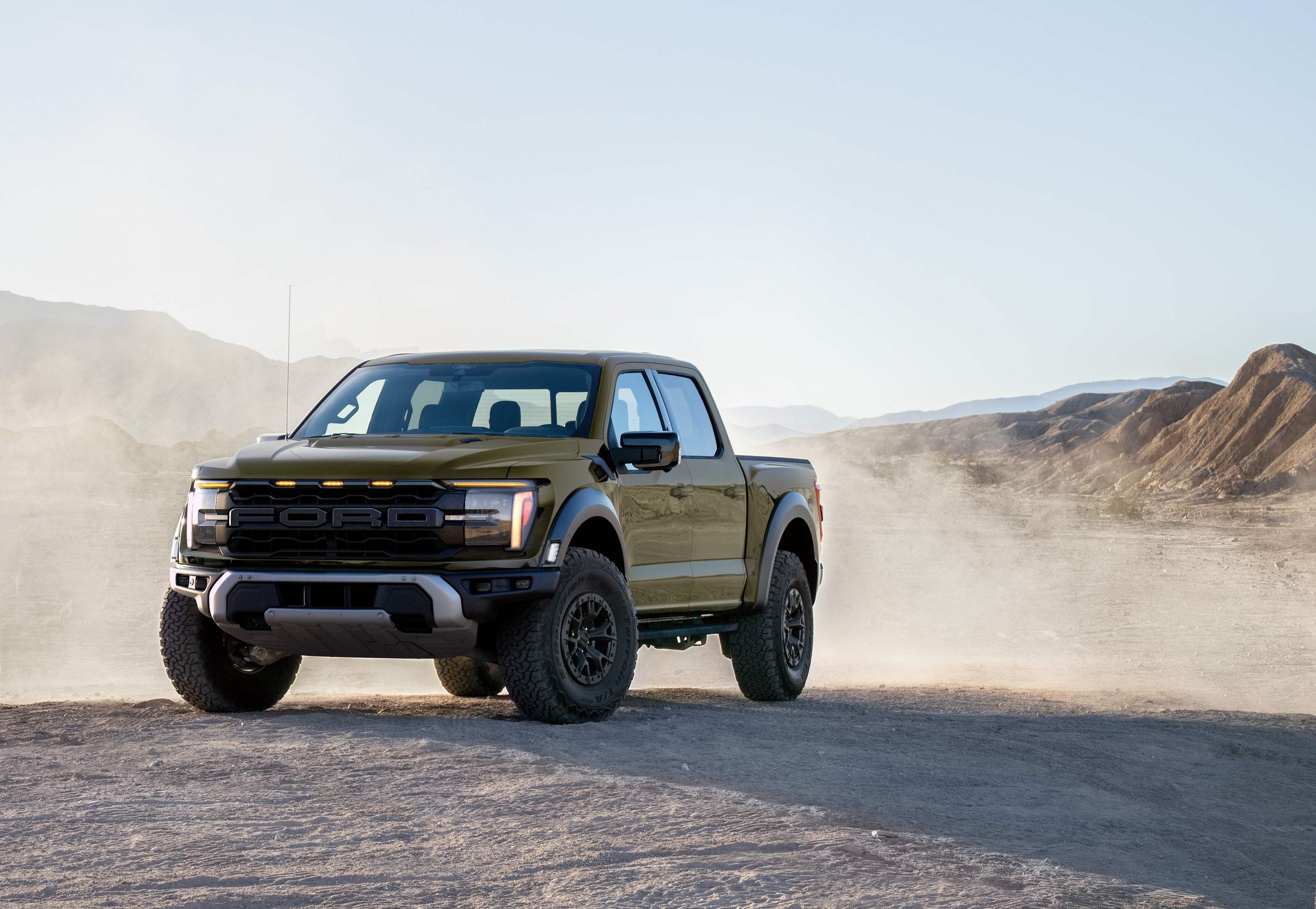 A dark olive-green 2026 Ford F-150 Raptor SuperCrew on a dusty desert road, seen from the front three-quarter with hills behind