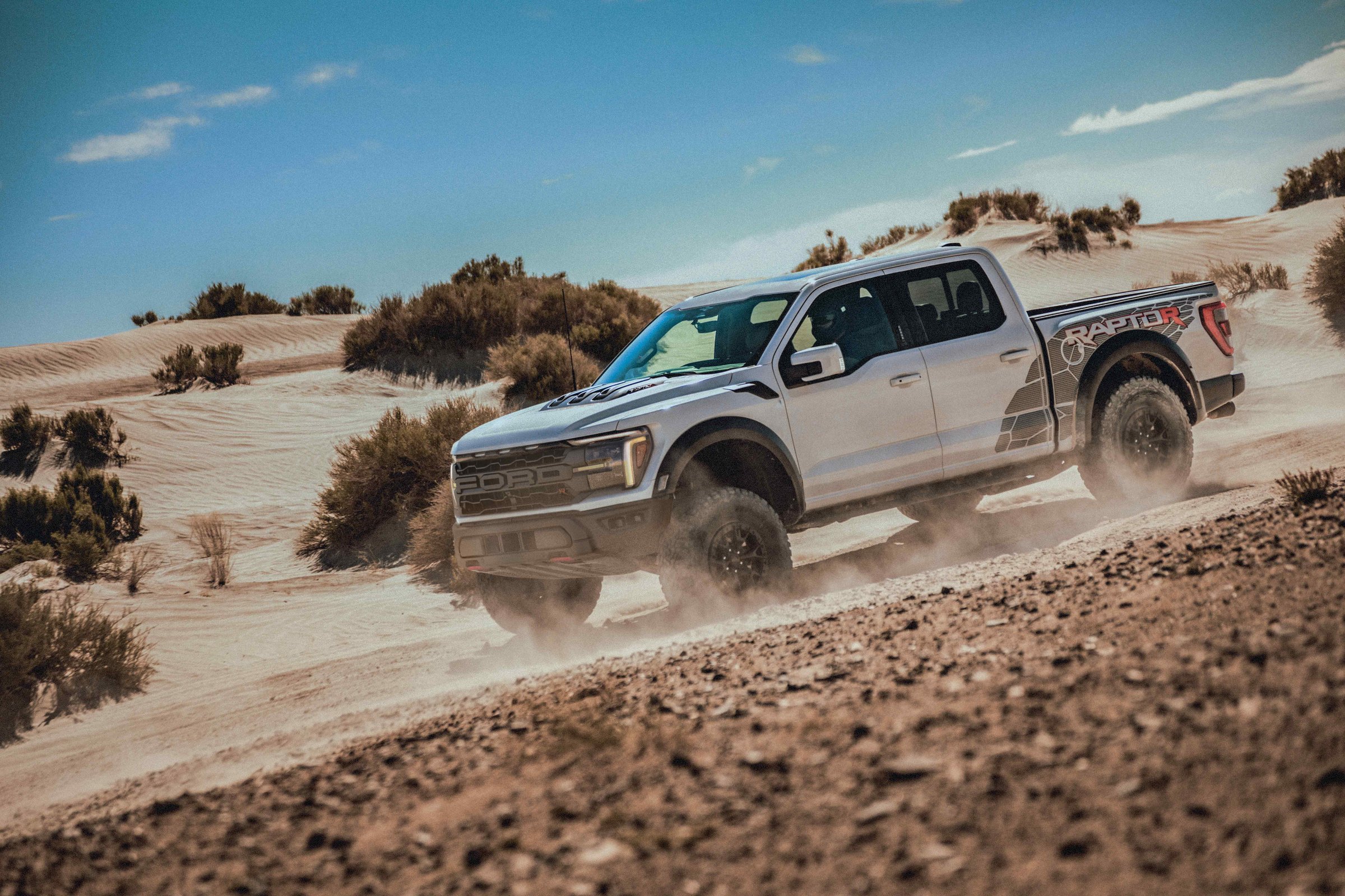 A white Ford F-150 Raptor R with Raptor R bed graphics climbing a sand dune at speed, throwing up dust