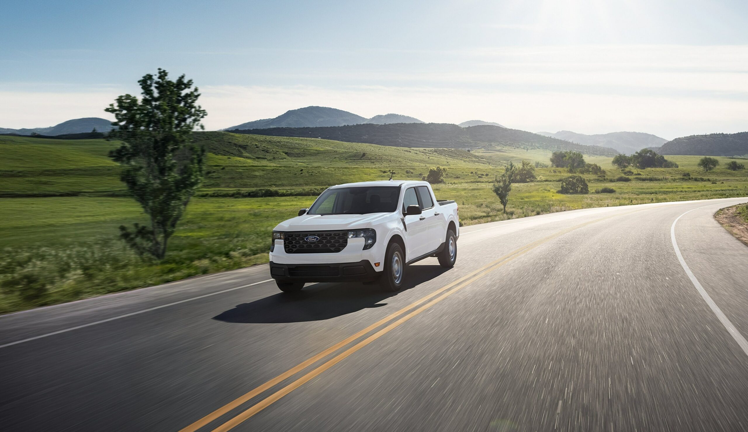 A white 2026 Ford Maverick driving on a two-lane road through green rolling hills
