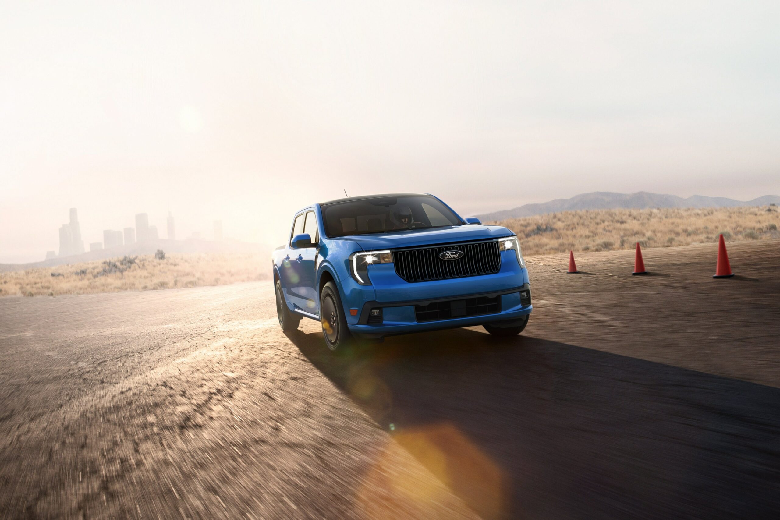 A blue 2026 Ford Maverick Lobo cornering between traffic cones on a closed course at dusk, city skyline behind