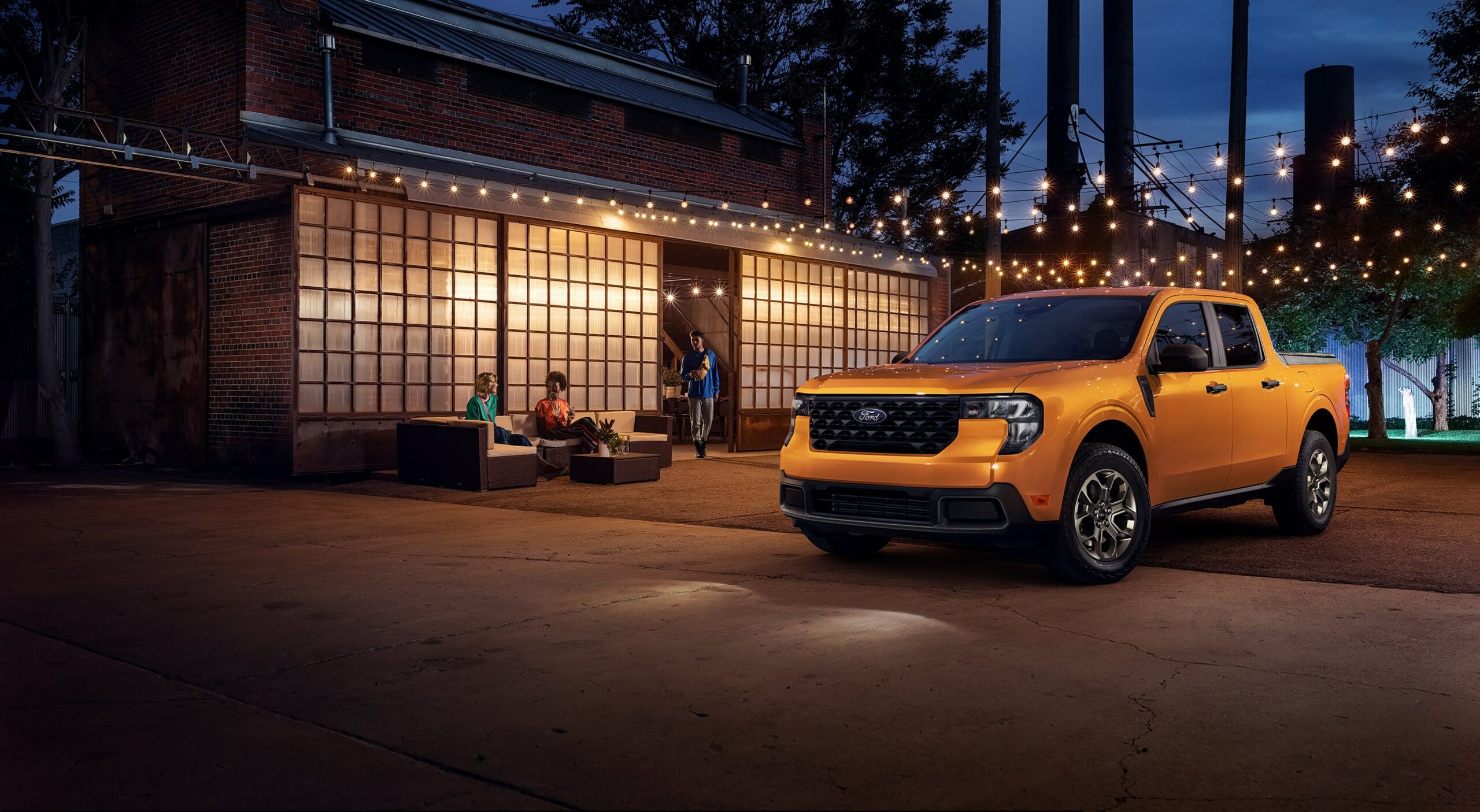 An orange 2026 Ford Maverick parked at night outside a lit venue strung with cafe lights