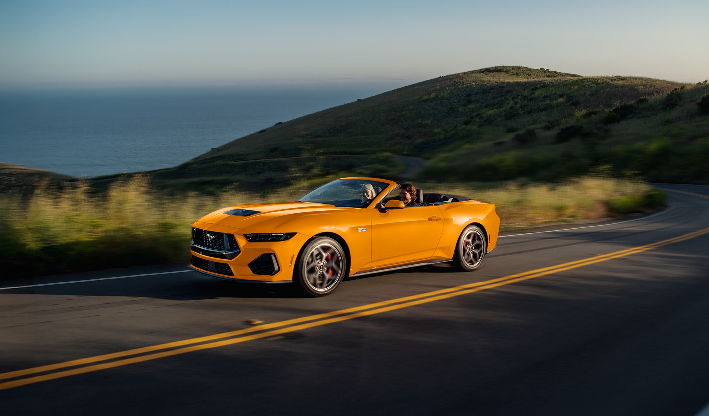 An orange 2026 Ford Mustang GT Premium Convertible with the roof down on a two-lane coastal road above the ocean