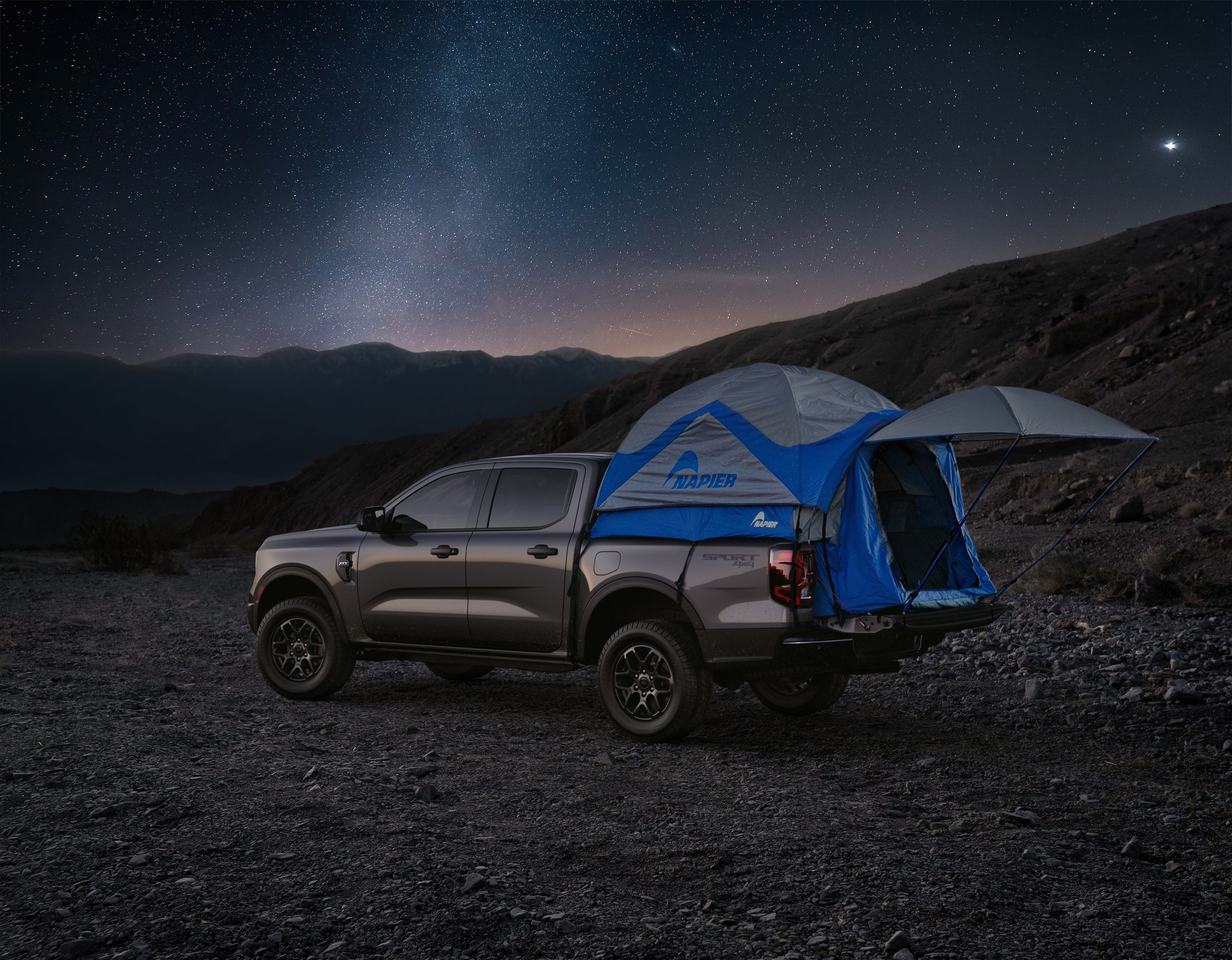 A carbonized gray 2026 Ford Ranger parked on a rocky hillside under a starry night sky, with a blue tent pitched in its cargo box and an awning extended