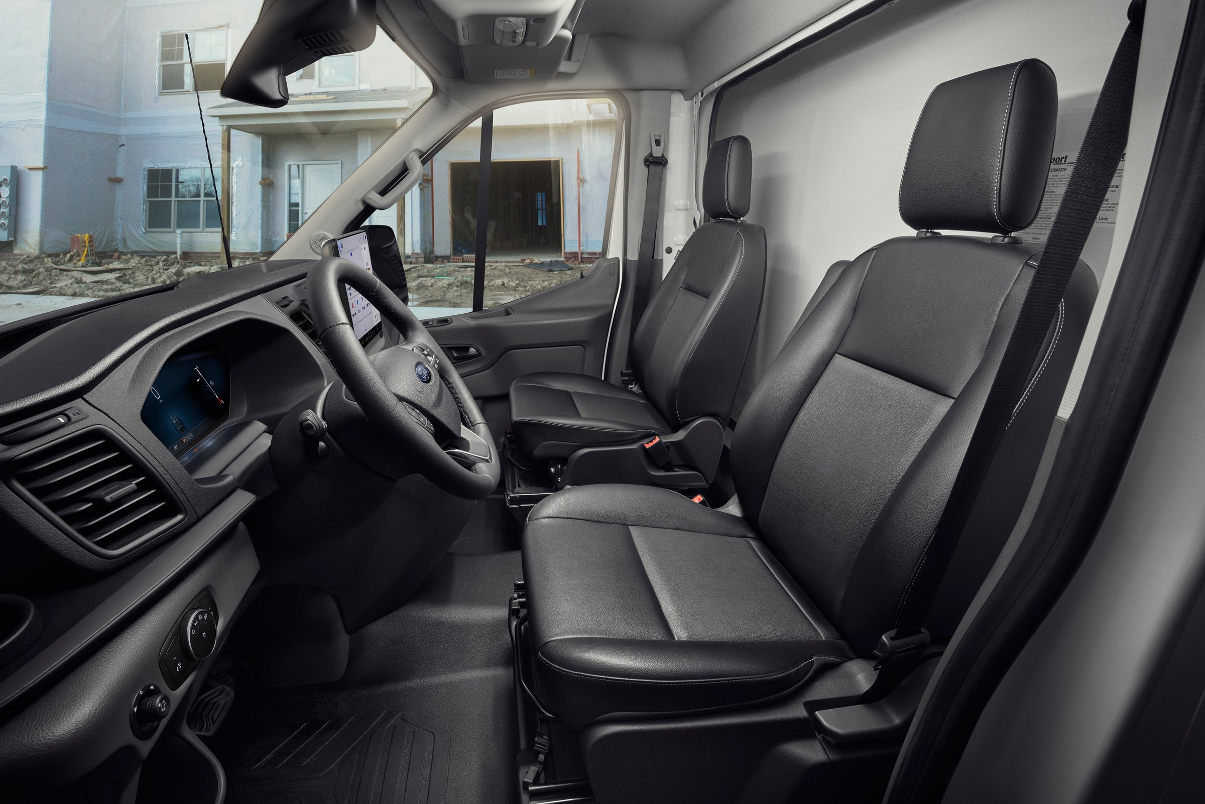 The front cab of a Ford Transit seen from the passenger side, with black cloth seats, the steering wheel and the center touchscreen