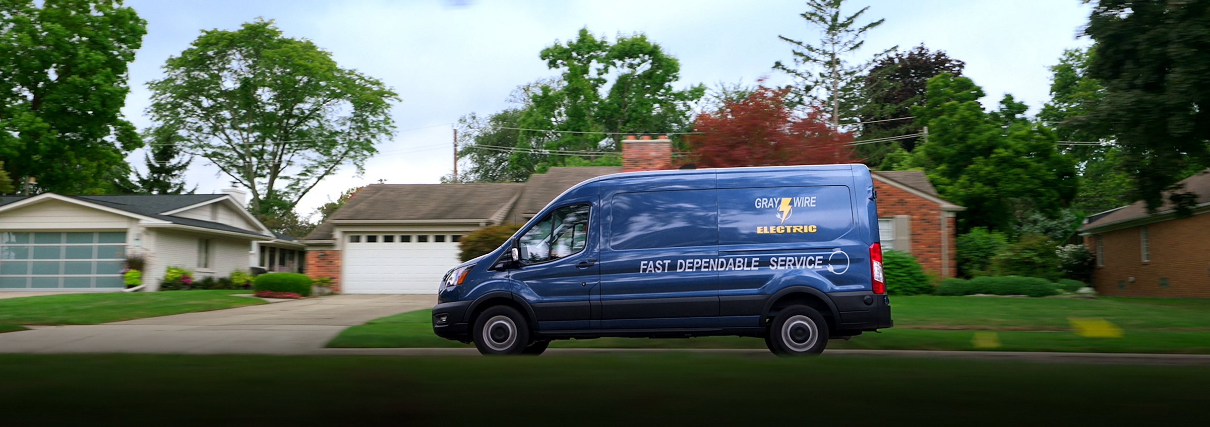 A dark blue Ford Transit cargo van in fleet livery photographed side-on in front of a house, showing its full medium-roof profile