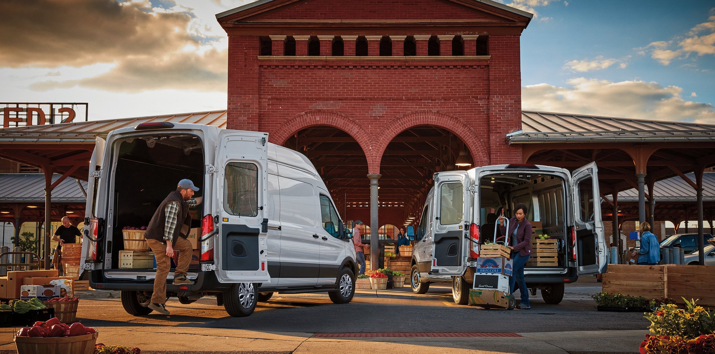 Two white high-roof Ford Transit cargo vans backed up to a brick market building with their rear doors open, workers wheeling crates out on hand trucks